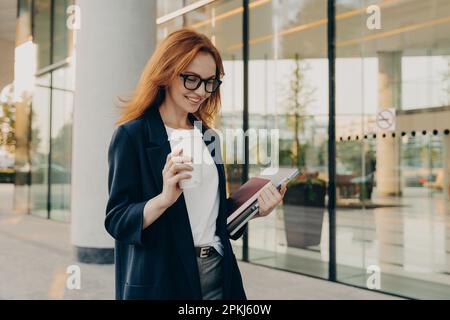 Souriante femme d'affaires vêtue de vêtements formels boissons café porte un carnet pour faire des notes ordinateur portable focalisé vers le bas avec Happy expres Banque D'Images