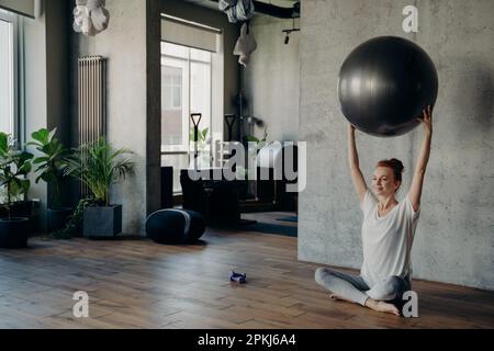 Jeune femme souriante aux cheveux rouges s'exerçant dans un studio de fitness, tenant une grande boule d'exercice argentée avec les deux mains au-dessus de sa tête et appréciant Li sain Banque D'Images