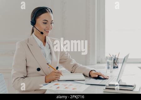 Vue latérale d'une jeune femme d'affaires souriante prenant part à une conférence Web, regardant l'écran d'un ordinateur portable, opérateur de centre d'appels consultant le client wh Banque D'Images