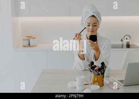 Jeune femme rafraîchie applique de la poudre pour le visage avec une brosse cosmétique se regarde dans le miroir pose à la cuisine à la maison s'assoit au bureau avec des produits de beauté wea Banque D'Images