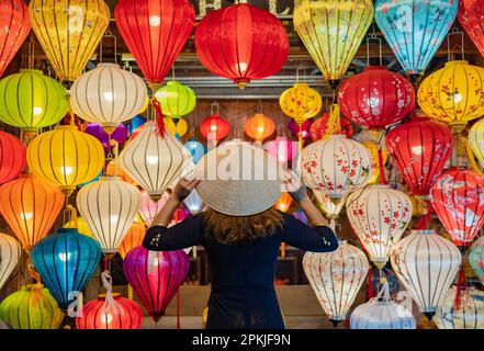 Vue arrière anonyme femme en robe touchant le chapeau conique asiatique sur la tête tout en se tenant près de lanternes colorées accrochées au plafond Banque D'Images