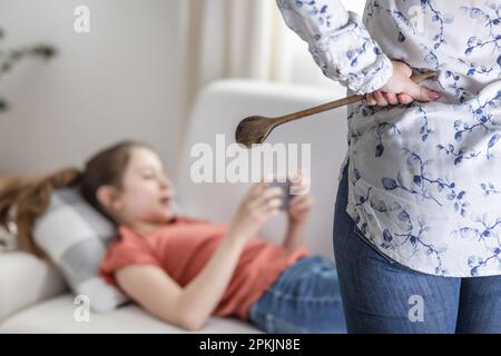 Une adolescente ignore que sa mère se tient contrariée avec une cuillère en bois à côté d'elle et continue à être sur son téléphone mobile. Banque D'Images