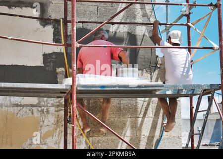 Deux constructeurs se sont assis sur un échafaudage à l'extérieur d'un bâtiment pour travailler sur le plâtrage d'un mur Banque D'Images