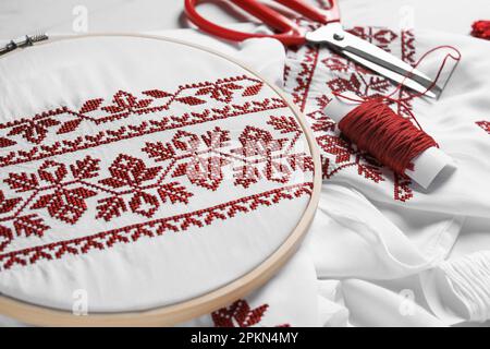 Chemise avec motif brodé rouge en cerceau, ciseaux et fil sur table, gros plan. Vêtements nationaux ukrainiens Banque D'Images