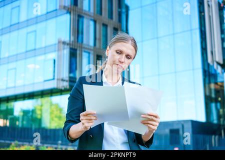 Élégante femme d'affaires caucasienne en tenue formelle tient des documents en papier dans ses mains sur le fond d'un immeuble de bureaux, un employé de bureau près du centre d'affaires. Banque D'Images