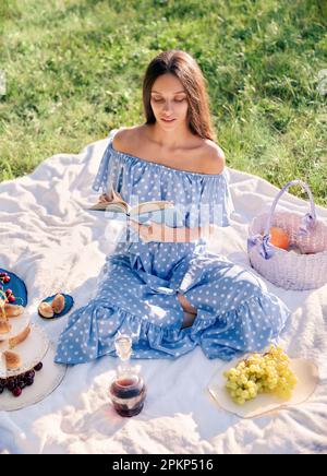 Scène de pique-nique: Jolie fille assise sur un écossais et livre de lecture dans le parc. Portrait extérieur de la belle jeune femme en robe bleue Banque D'Images