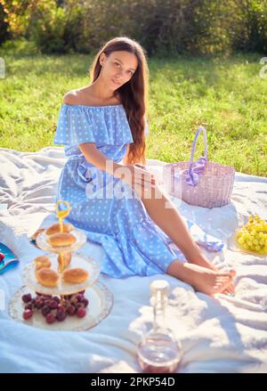 Scène de pique-nique : petite fille douce assise sur un terrain de pique-nique dans le parc. Portrait extérieur de jeune femme adorable en robe bleue Banque D'Images