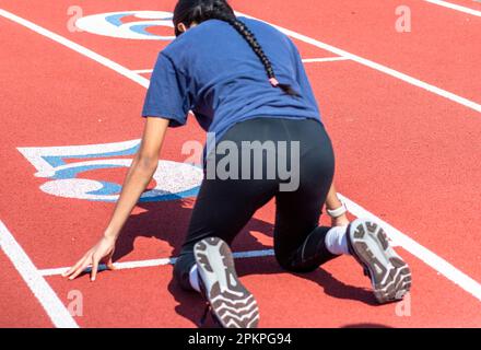 Vue arrière d'une fille d'école secondaire s'exerçant à démarrer une course dans la rangée cinq sur une piste portant le spandex noir et un t-shirt. Banque D'Images