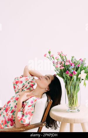 Jeune femme rêveuse assise dans un fauteuil avec des yeux fermés à côté d'un vase avec un beau bouquet de fleurs sur la table. Fête des mères, anniversaire, anniversaire Banque D'Images