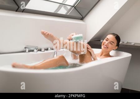 Femme attrayante prenant le pied de lavage de bain avec éponge dans la salle de bains Banque D'Images