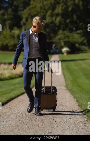 Un homme en costume bleu marche avec une valise sur un chemin de gravier Banque D'Images