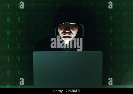 Hacker dans le sweat à capuche assis à l'avant de son ordinateur portable la nuit, les chiffres verts sur l'arrière-plan. Cybercriminel Banque D'Images