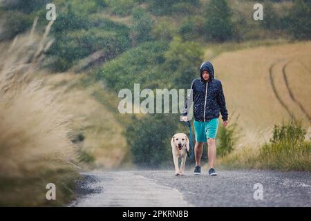 Homme avec un chien en laisse marchant ensemble sur une route rurale humide par forte pluie. Le propriétaire d'un animal de compagnie et son labrador Retriver par mauvais temps. Banque D'Images