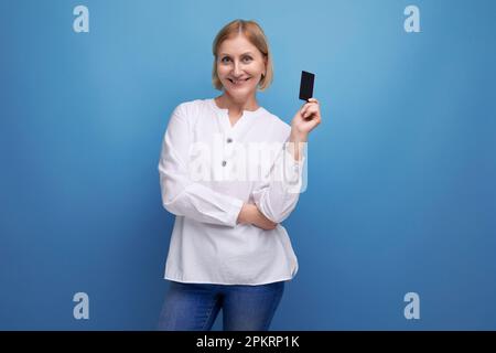 femme d'âge moyen cintrée et pensive blondie dans un chemisier blanc tenant une carte en plastique pour faire du shopping Banque D'Images