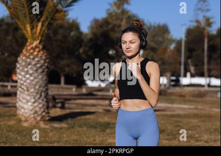 une jeune femme mince en tenue de sport écoute de la musique, balado sur un casque pendant qu'elle court à l'extérieur au parc avec des palmiers. Entraînement pendant les vacances d'été, Banque D'Images