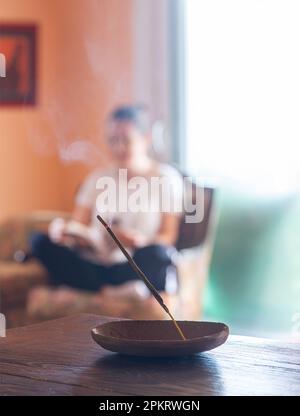 Bâton d'Encens brûlant avec une femme assise crosslegged lisant un livre en arrière-plan dans une chambre confortable avec des murs orange Banque D'Images