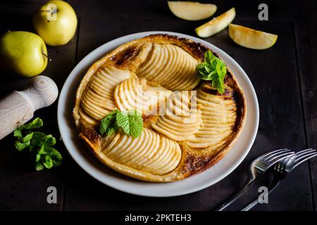 Tarte française aux pommes : tentation sur fond noir. Tarte traditionnelle ouverte avec fines tranches de pommes, décorée de feuilles de menthe. Banque D'Images