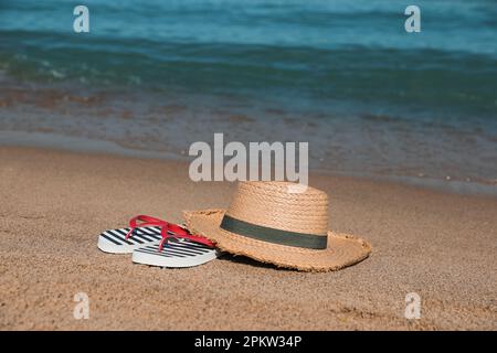 Chapeau de paille élégant et tongs rayés sur la plage de sable près de la mer Banque D'Images
