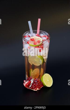 Un grand verre à facettes de jus d'agrumes avec glace, graines et pailles sur fond noir, à côté de morceaux de lime mûre et d'un fruit de grenade cassé. ver Banque D'Images