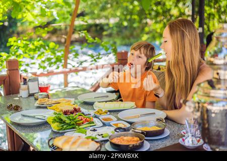 Maman et son prennent un petit déjeuner turc. Table de petit-déjeuner turc. Pâtisseries, légumes, légumes verts, olives, fromages, œufs frits, épices, confitures, miel, thé Banque D'Images