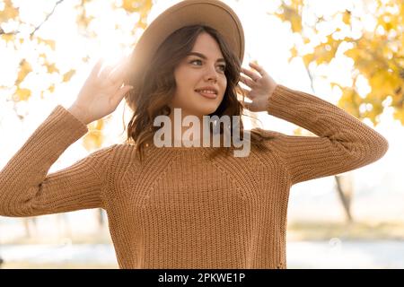 Belle femme élégante tenant un chapeau sur la tête, marchant dans le parc, vêtue d'un pull chaud. Jeune femme aux cheveux marron clair souriant par temps chaud Banque D'Images