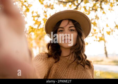 Jolie jeune fille hipster dans un chapeau élégant dans un pull tendance debout dans le parc et fait un selfie sur un téléphone. Prendre soi-même Banque D'Images