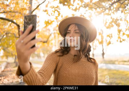 Belle dame rêveuse faire des selfies portant un chapeau marchant dans le parc d'automne, appréciant la journée chaude. Copier l'espace Banque D'Images