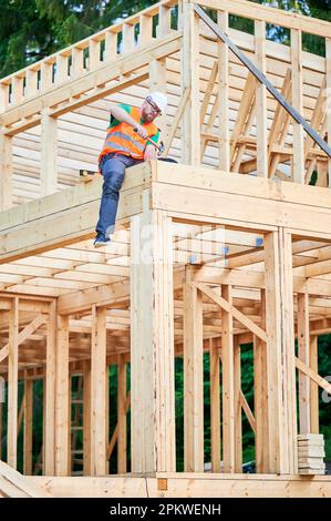 Carpenter construisant une maison en bois de deux étages. Un homme barbu fait marteler des ongles dans la structure tout en portant un casque de protection et un gilet de construction. Concept de construction écologique moderne. Banque D'Images