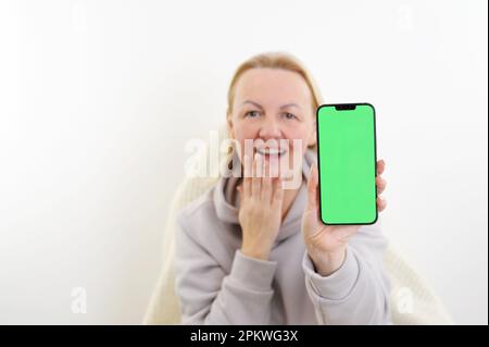 Une jeune femme au visage surprise pointe un doigt vers un smartphone avec un écran vert dans sa main. Photo de haute qualité Banque D'Images