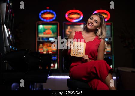 Portrait d'une jeune fille de race blanche belle et bien habillée en robe rouge jouant dans le Casino et célébrez la tenue de la carte de jeu de bois de Bingo Banque D'Images