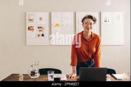 Une jeune architecte d'intérieur souriant à la caméra tout en se tenant derrière son bureau. Bonne femme d'affaires avec lunettes de vue travaillant avec un ordinateur portable Banque D'Images
