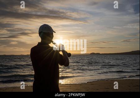 Silhouette d'un musicien jouant de la clarinette sur la plage de Milford au lever du soleil. Le soleil brille à travers les nuages. Auckland. Banque D'Images