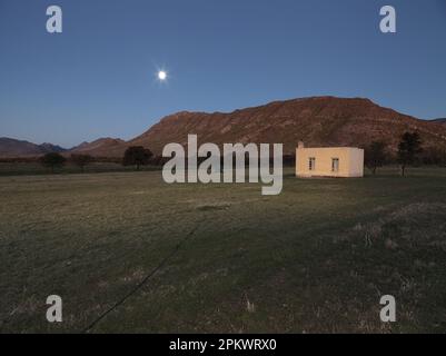 La pleine lune s'élève au-dessus de ou Kloofberg, une petite montagne au pied de la chaîne de montagnes Swartberg. Petite maison de travail à la périphérie de Banque D'Images