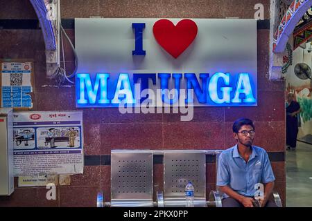 Un passager mâle est assis sur un banc à la gare de Matunga (Central Railways), Mumbai, Inde, sous un panneau lumineux au néon indiquant « I Love Matunga » Banque D'Images