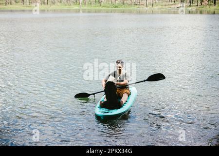 Homme et chien dans un kayak sarcelle sur un lac avec une forêt derrière eux. Banque D'Images