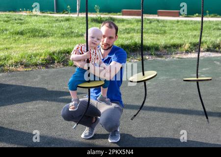 Papa tient son fils sur une balançoire ronde sur une corde dans l'aire de jeux Banque D'Images