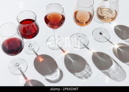 Verres à vin debout en ligne, remplis de différents vins rouges, blancs et roses sur fond blanc. Réflexion et ombres Banque D'Images