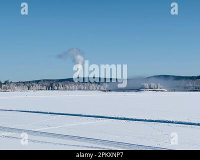 Paysage hivernal dans le nord de la Suède avec un lac gelé recouvert de neige et une cheminée industrielle lointaine émettant de la vapeur Banque D'Images