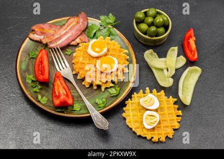 Gaufres, œufs frits, bacon et fourchette sur l'assiette. Olives dans un bol. Légumes hachés et gaufres sur la table. Vue de dessus. Arrière-plan noir. Banque D'Images