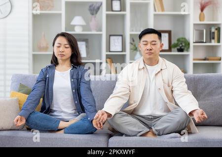 Une famille asiatique heureuse à la maison sur un canapé méditant, couple homme et femme assis en position lotus dans le salon avec les yeux fermés se reposant ensemble. Banque D'Images