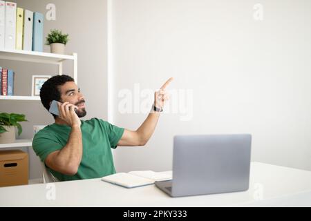 Homme arabe joyeux du millénaire avec des appels de barbe par téléphone, travaille avec un ordinateur portable sur le lieu de travail, pointe le doigt sur l'espace vide Banque D'Images