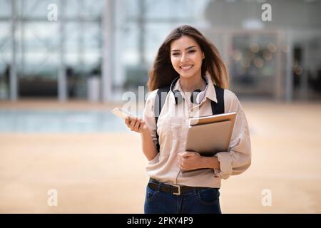 Bonne étudiante de brunette blanche millénaire avec sac à dos, livres et smartphone dans un casque sans fil Banque D'Images