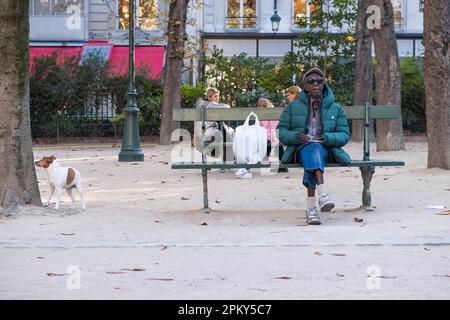 Un homme noir plus âgé profite d'un moment calme sur un banc de jardin, en prenant une pause des rues animées de la ville tout en appréciant une fumée. chien marche à côté de lui. Banque D'Images