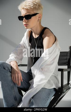 femme blonde dans des lunettes de soleil tendance et des vêtements élégants assis sur une chaise pliante sur le gris Banque D'Images