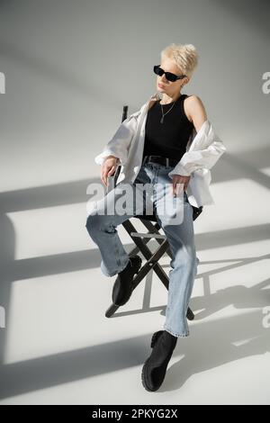 pleine longueur de femme blonde élégante en noir lunettes de soleil assis sur une chaise pliante sur le gris Banque D'Images