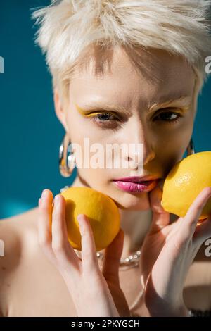 portrait d'une femme albino avec maquillage tendance et cheveux blonds posant avec des citrons mûrs sur le bleu Banque D'Images