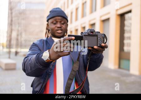 Joyeux jeune homme noir dans des vêtements tendance avec des tresses debout à l'extérieur et regardant l'appareil photo tout en prenant des photos sur un appareil photo professionnel Banque D'Images