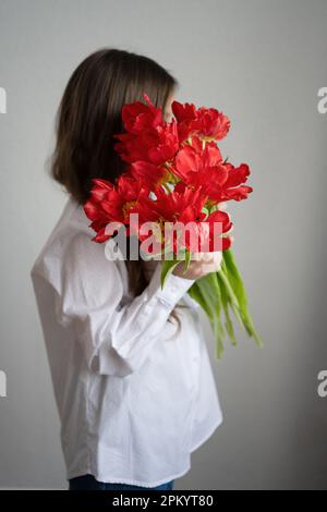 Anonyme jeune femme en chemise blanche tenant rouge Hippeastrum fleurs fleuries avec des tiges dans les mains et debout contre le mur gris Banque D'Images
