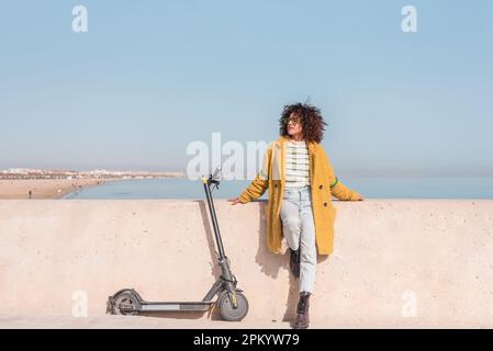 Une femelle rêveuse à poil chaud et des lunettes avec les yeux fermés, debout près d'un scooter électrique, au bord d'un remblai contre la mer Banque D'Images