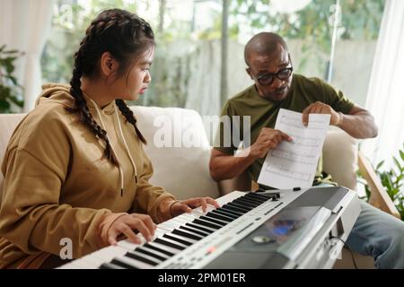 Fille regardant des notes de musique dans les mains de professeur et jouant le synthétiseur Banque D'Images